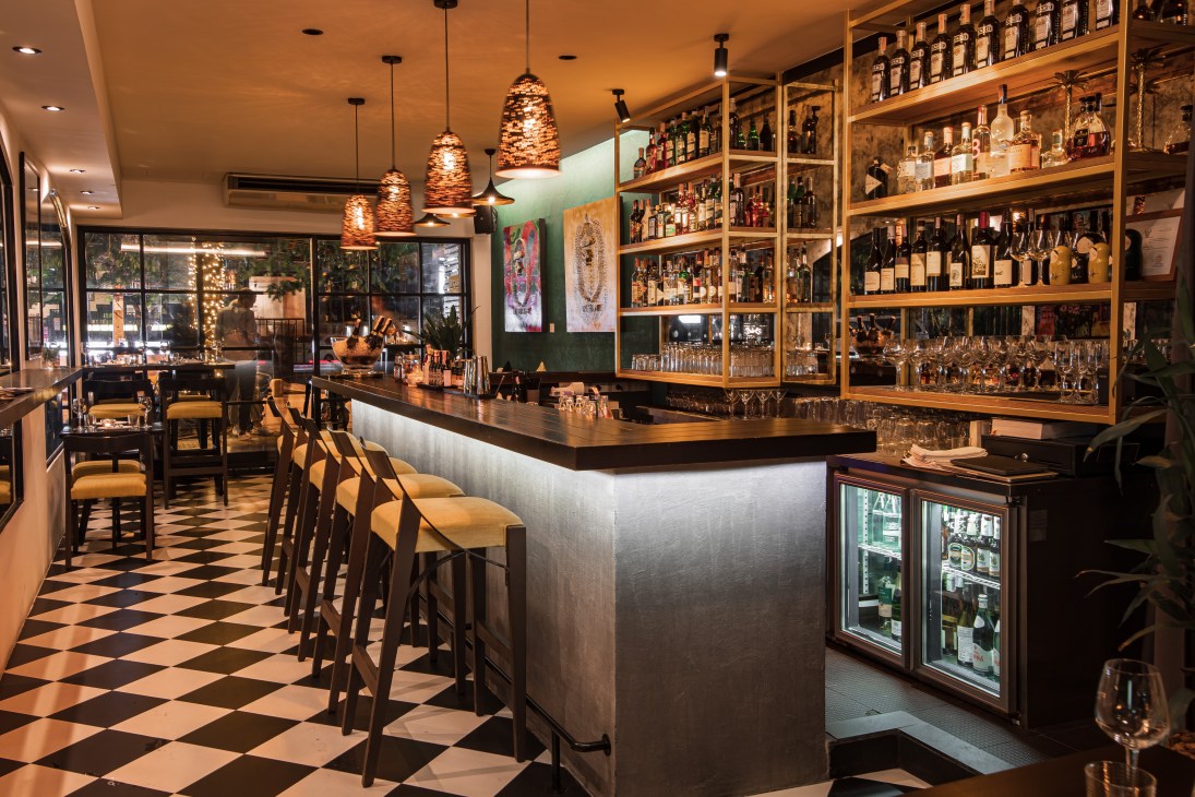 Interior of Bardo Social Bistro and Bar in Bangkok featuring a bar counter, shelving with bottles, pendant lighting, and checkered flooring.
