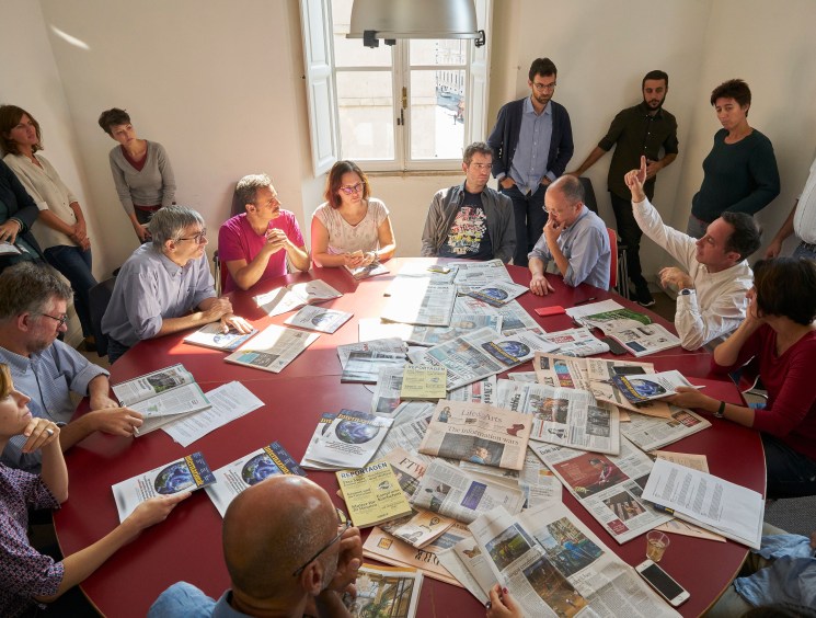 A group of people gathered around a large round table covered in newspapers and documents, engaged in discussion.