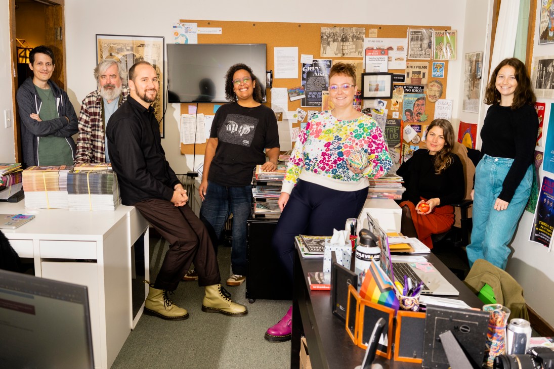 Matthew Steele and Little Village magazine team members in their Iowa City office with cork board and magazine covers on wall.
