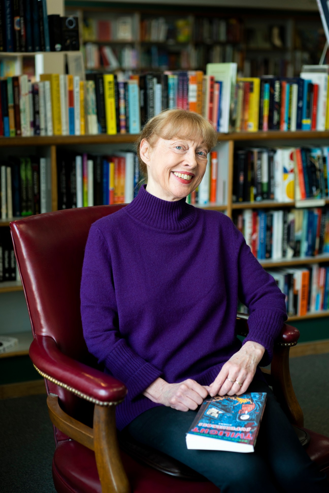 Jan Weissmiller, co-owner of Prairie Lights bookshop, sits in a burgundy leather chair holding a book, with bookshelves behind her.