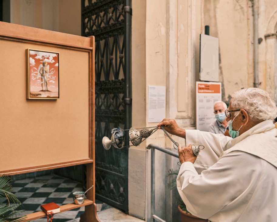 Elderly man in white robes examining an artwork while another masked person watches in background