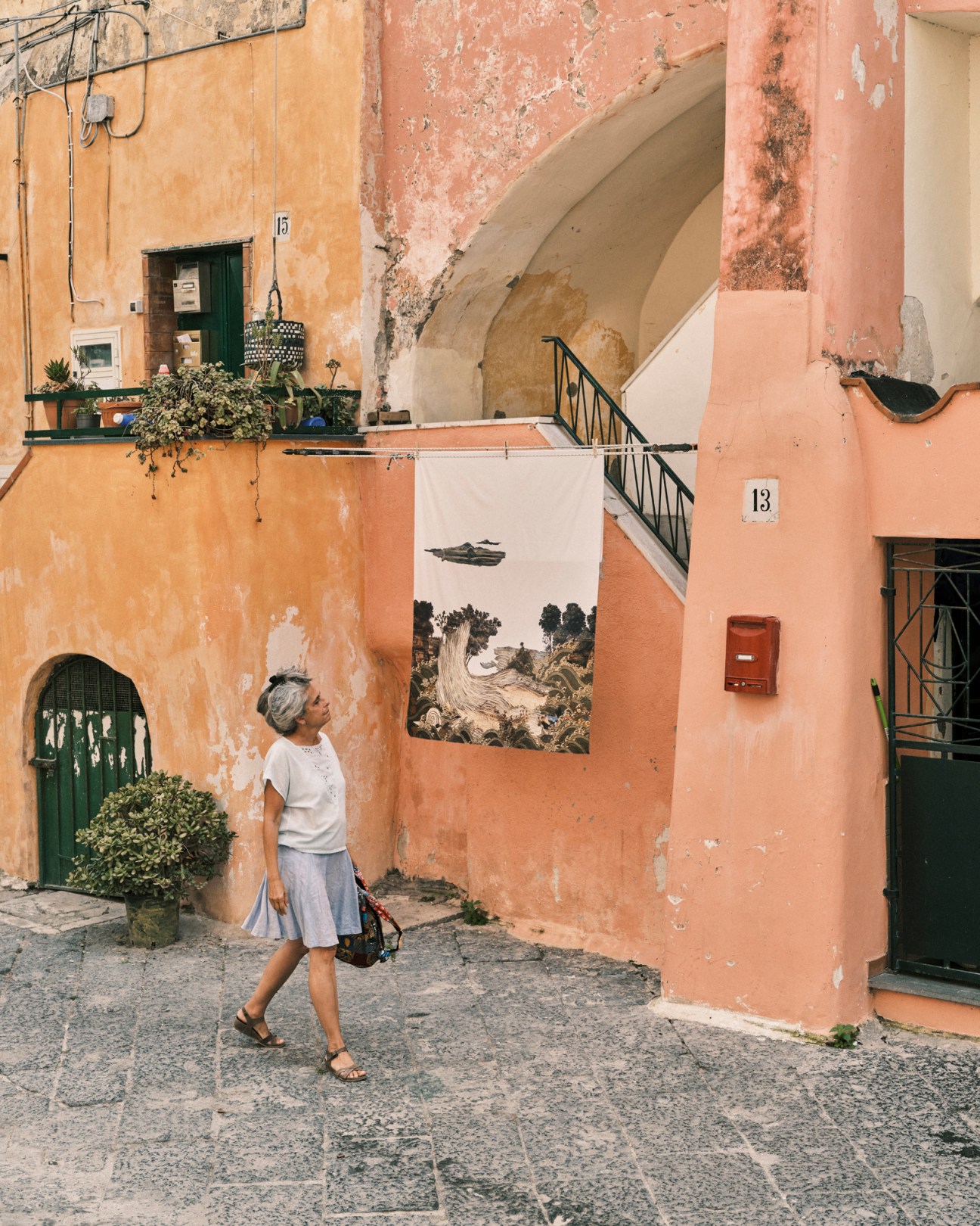 Woman in white shirt and light skirt walking past weathered pink and orange building walls with artwork display