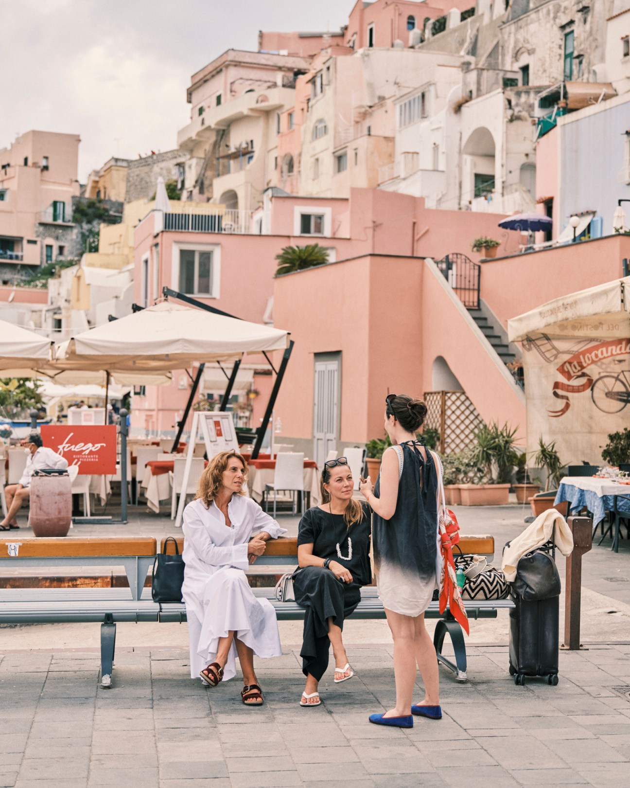 Three women sitting and standing on a bench in a piazza with Mediterranean architecture and outdoor dining in background