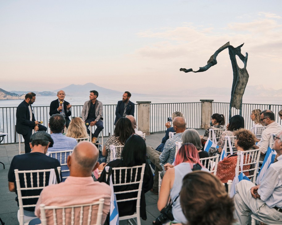 Panel discussion at outdoor art event with sculpture overlooking Mediterranean coastline at sunset