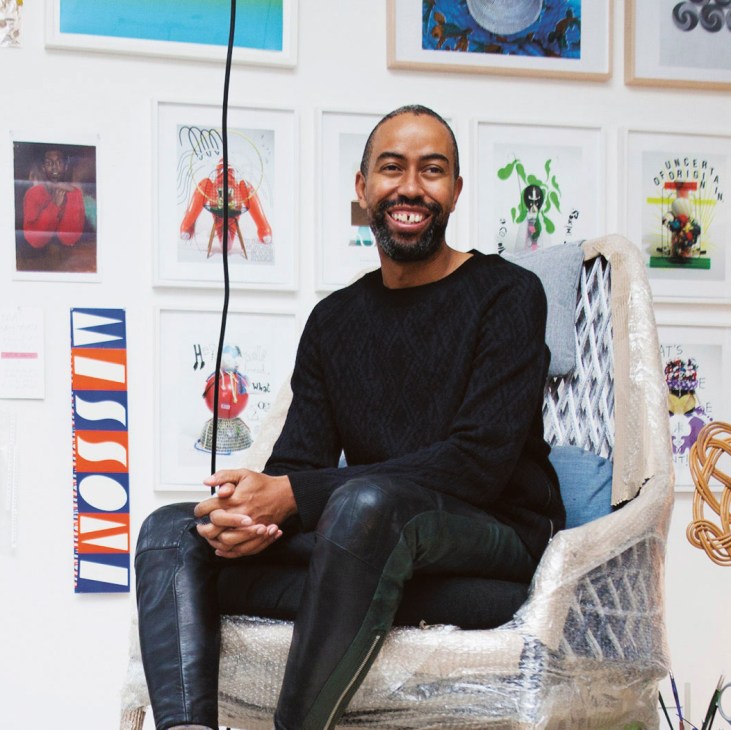 Stephen Burks, an African American designer, sits in a chair surrounded by artwork and design pieces in his studio.