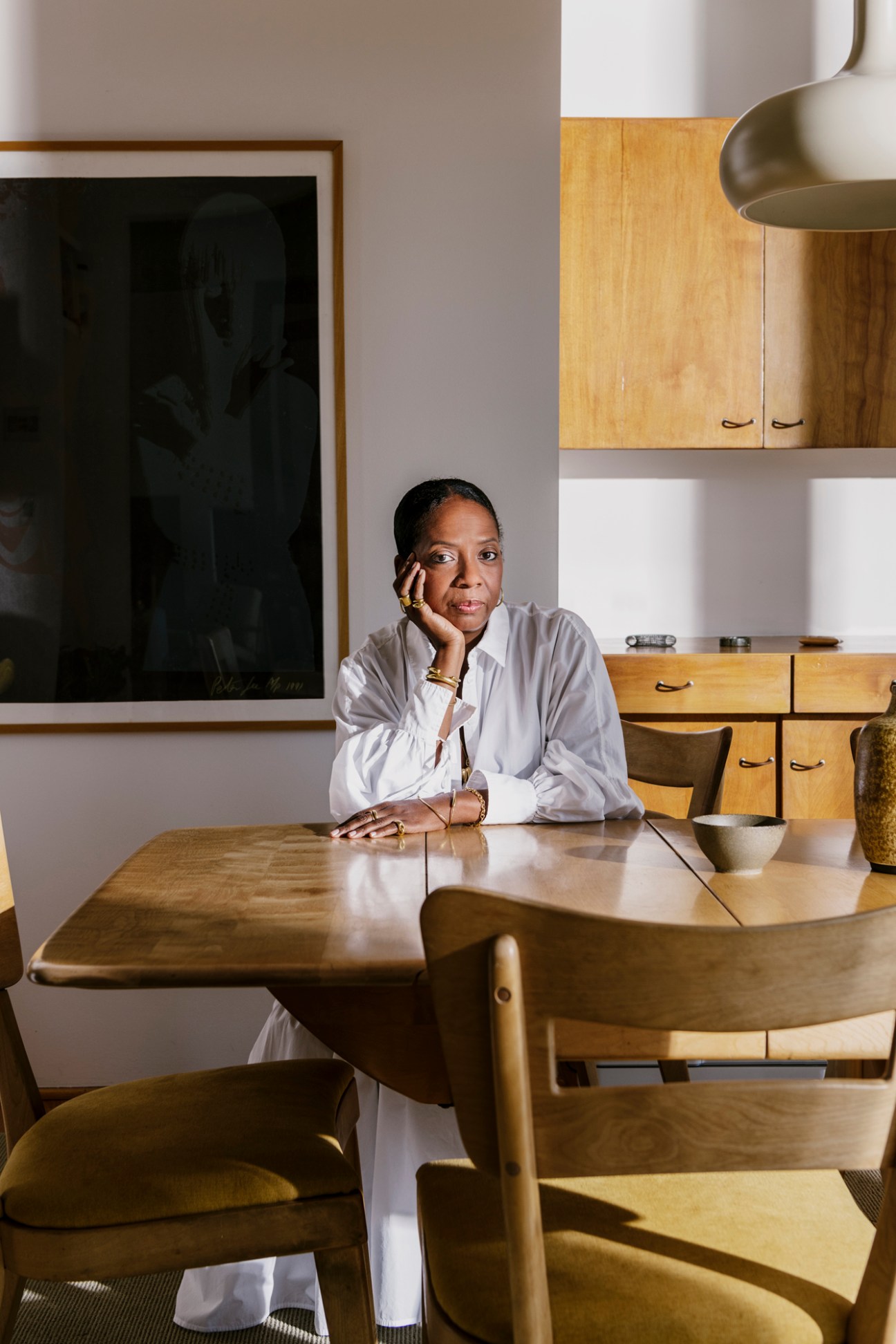 Jacqueline Rabun sits at a wooden table in her Los Angeles studio wearing a white shirt