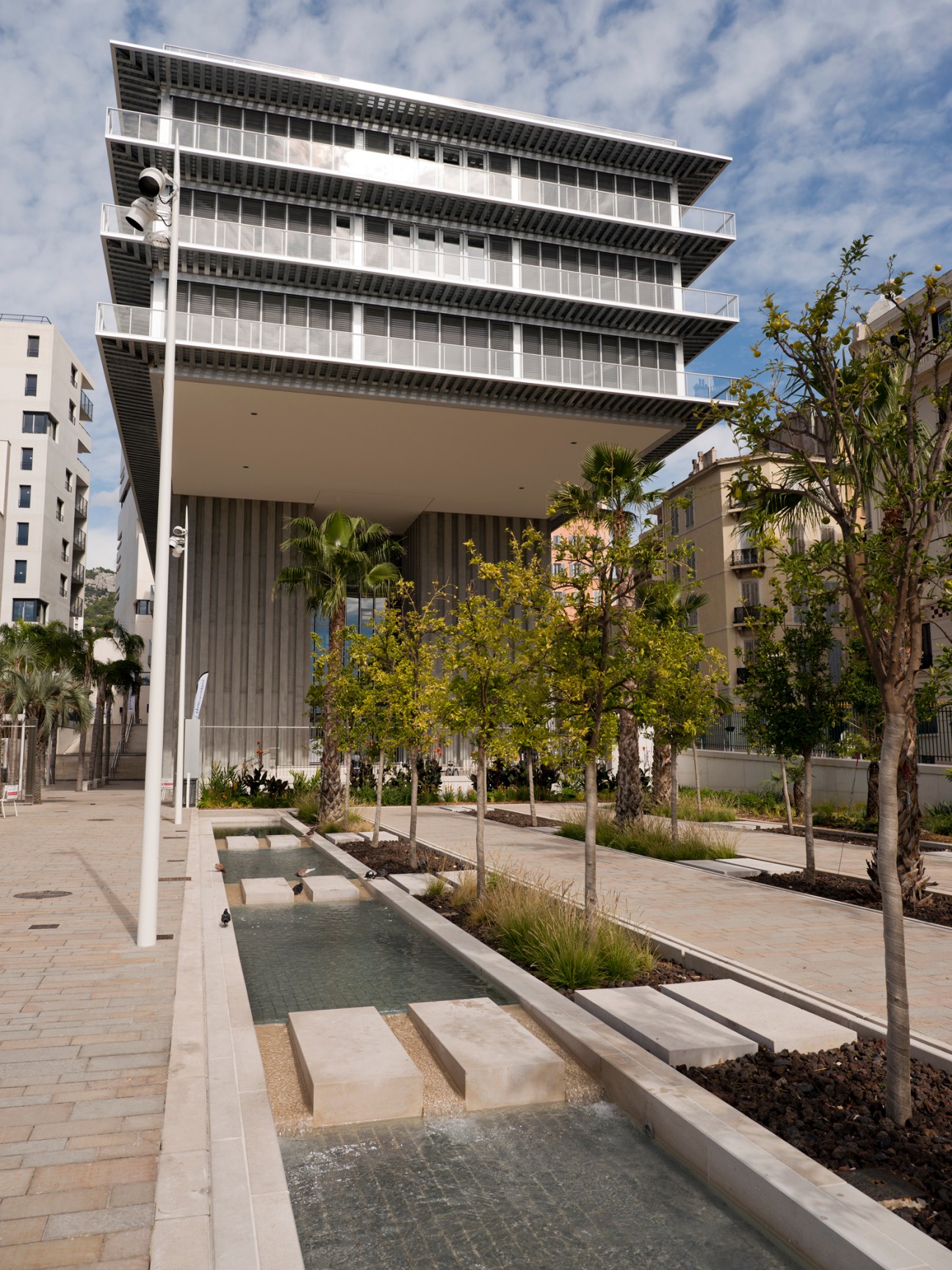 Modern white building with stacked horizontal levels and large windows, surrounded by palm trees and water channels in courtyard.