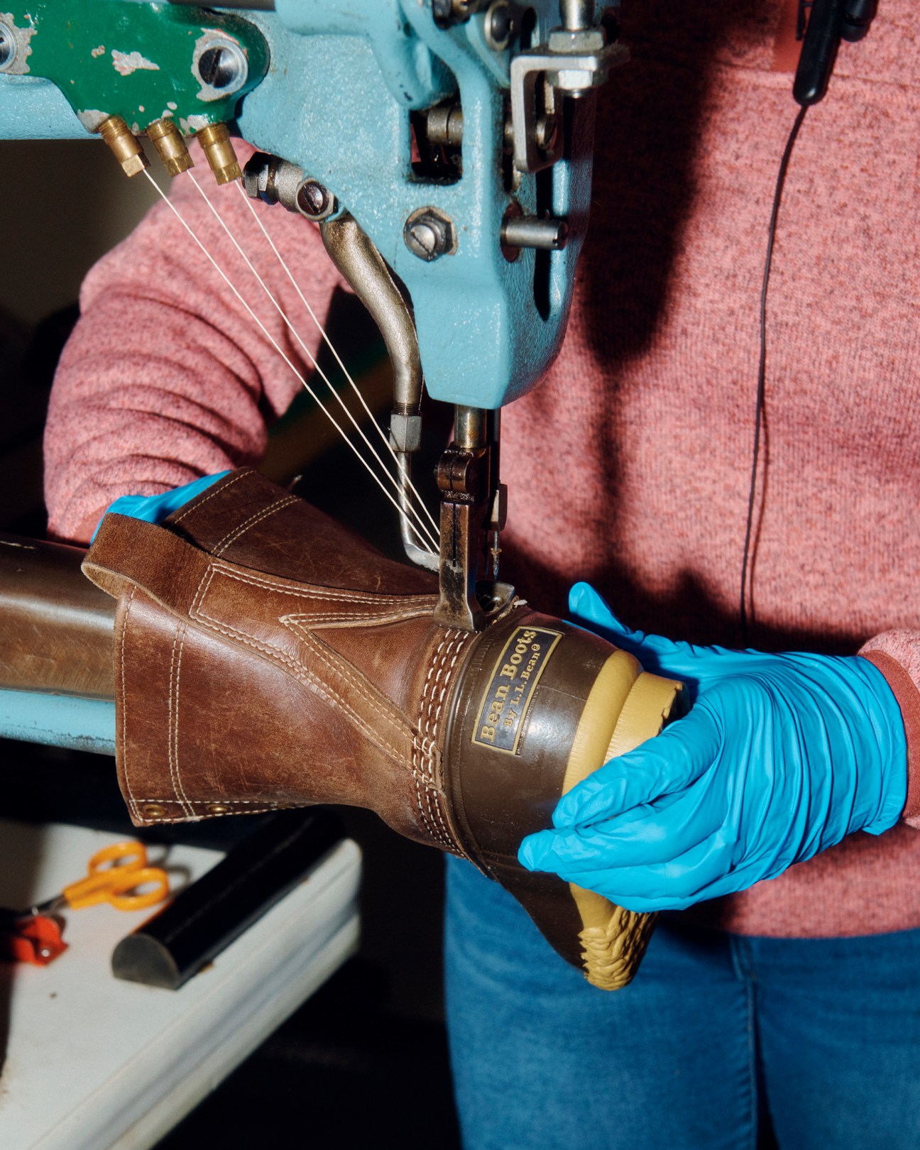 Close-up of hands in blue gloves operating a sewing machine to stitch brown leather Bean Boot showing signature triple stitch