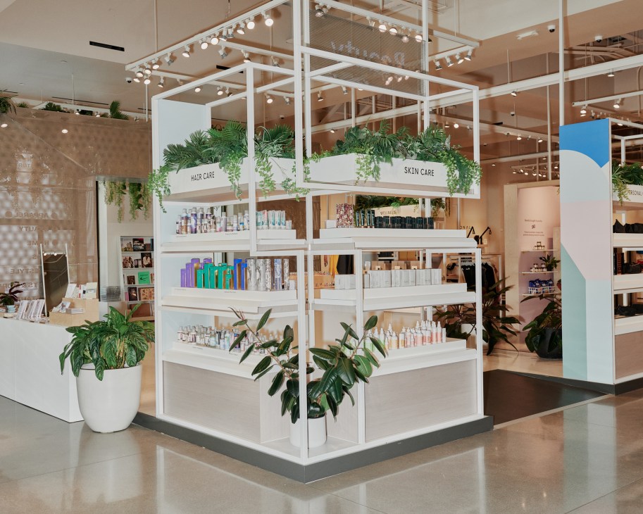 Interior of Neighborhood Goods department store showing a modern white shelving unit with hair care and skin care products, decorative plants, and track lighting.