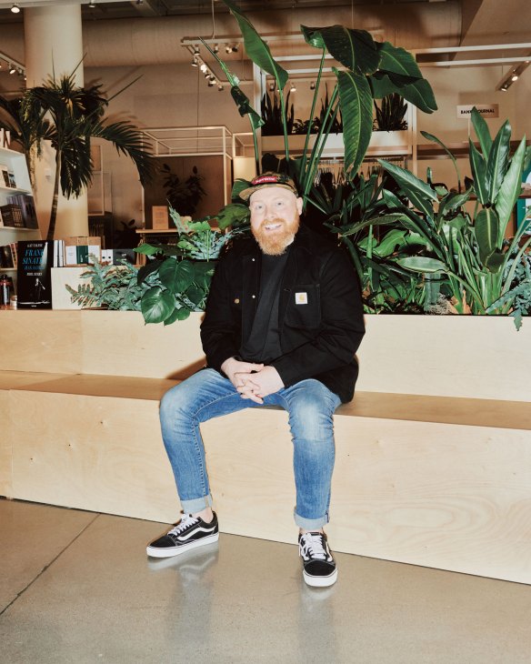 Matt Alexander sits on a wooden counter in Neighborhood Goods retail space with plants behind him.