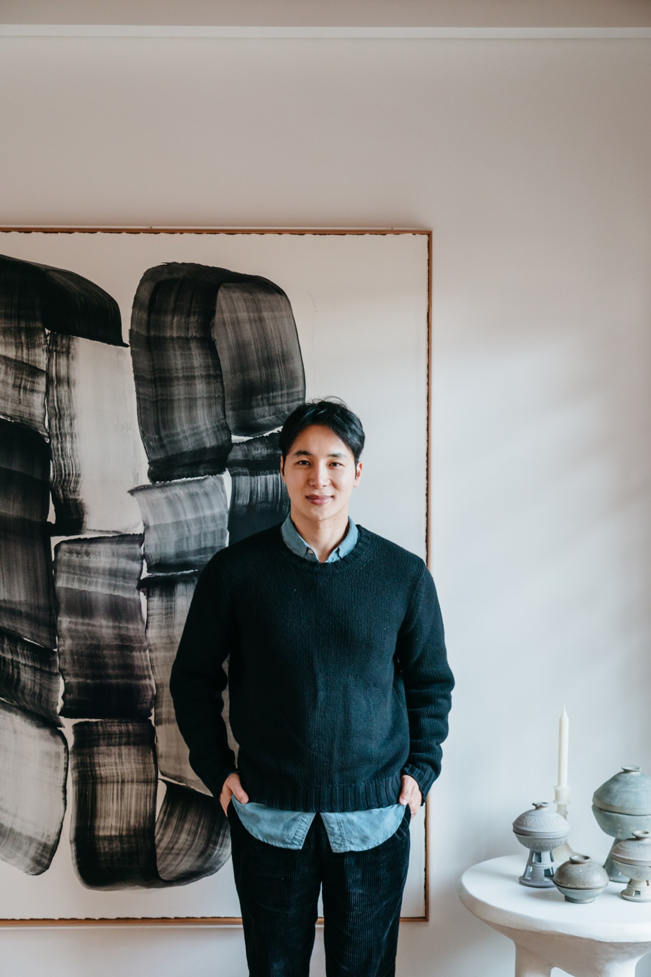 Teo Yang standing in front of abstract black brushstroke artwork with ceramic objects on white table
