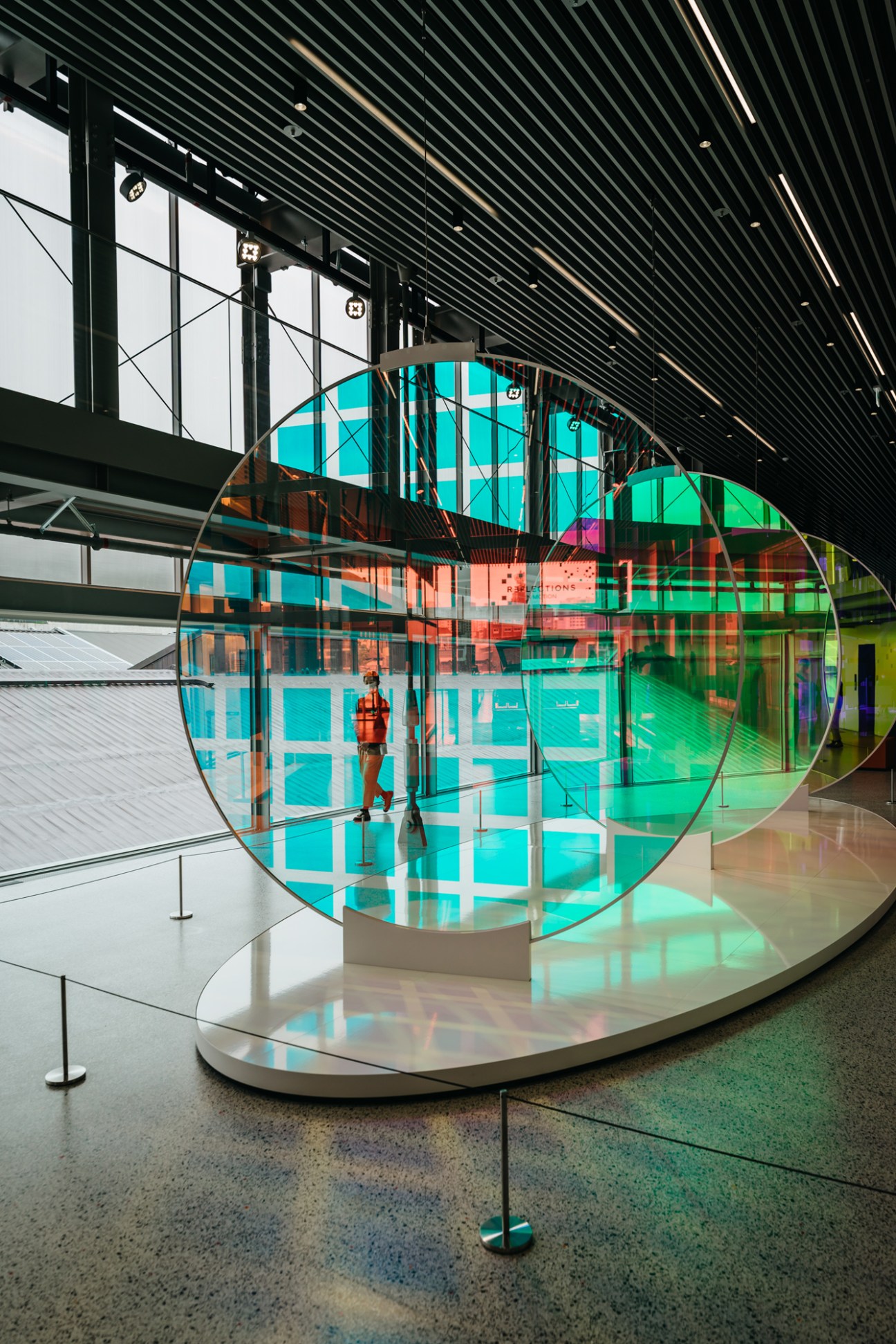 Colorful circular light installation with turquoise and green panels in a modern gallery space with industrial ceiling.
