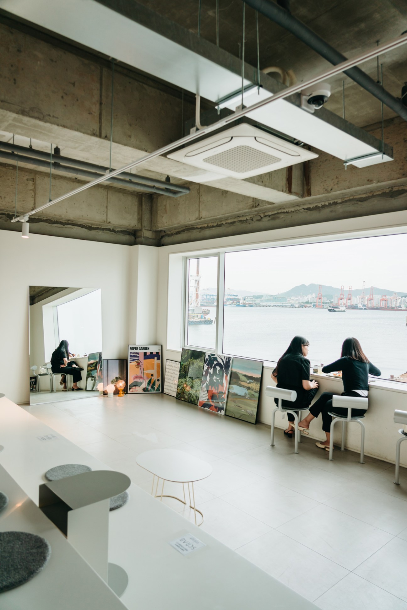 Modern gallery interior with visitors viewing artworks, industrial ceiling, and harbor view through large windows.