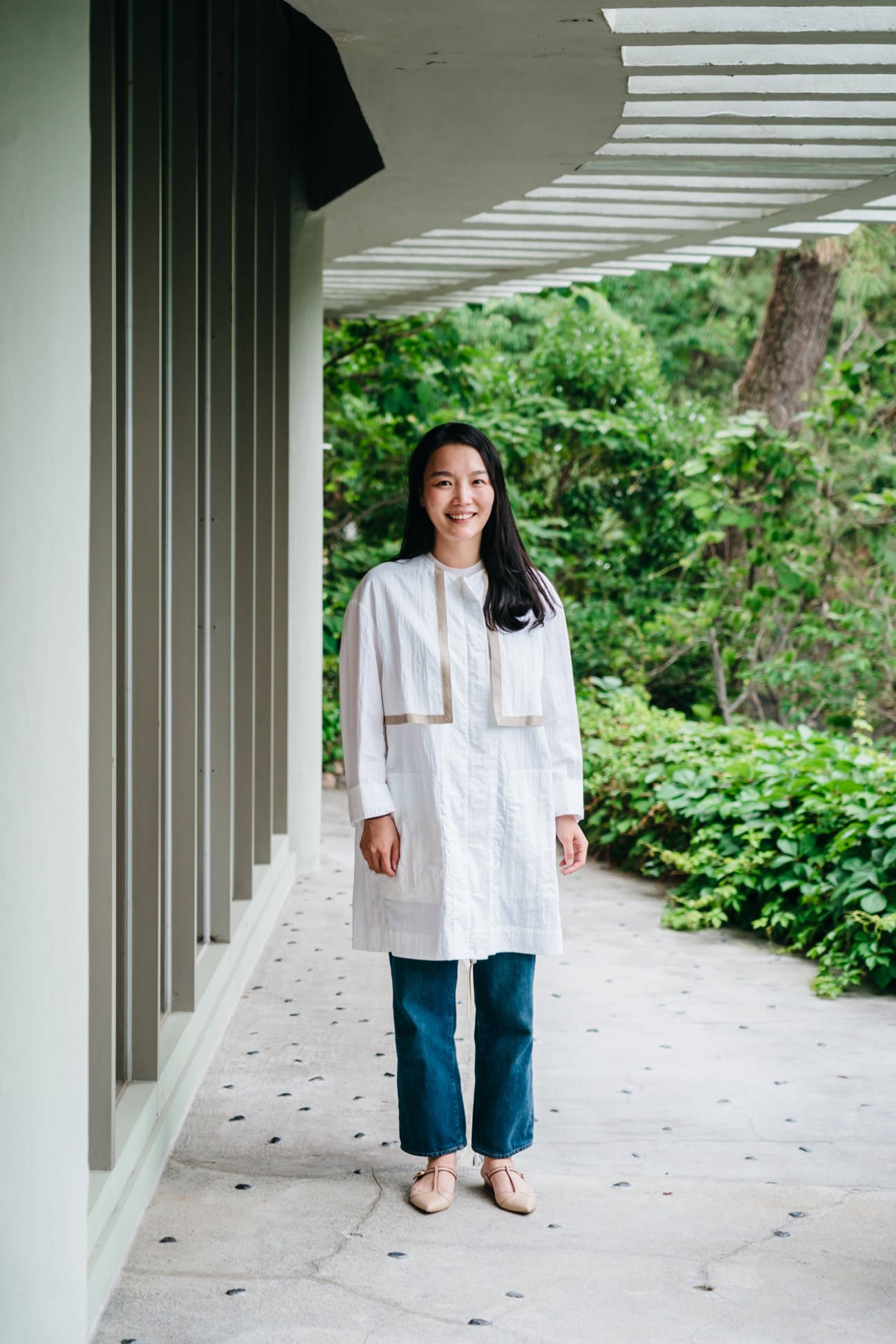 Woman in white shirt standing under modern covered walkway with green foliage in background.