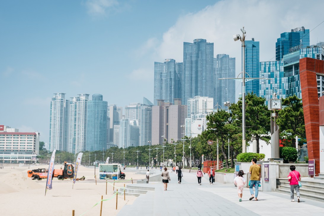Busan's beachfront promenade with sandy beach, pedestrians, and modern high-rise buildings skyline.