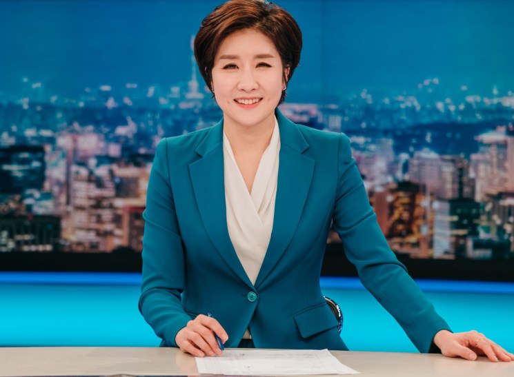 Lee So Jeong sits at a news desk in the KBS studio wearing a teal blazer and smiling at the camera