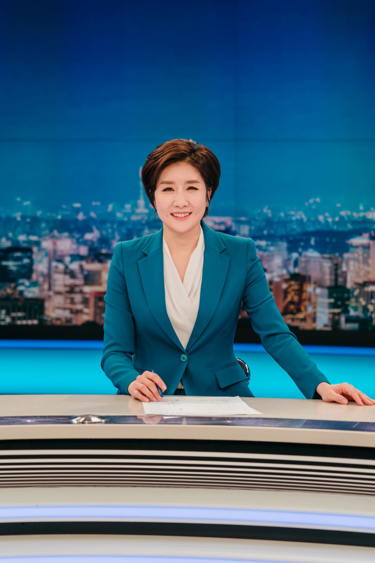 Lee So Jeong sits at a news desk in the KBS studio wearing a teal blazer and smiling at the camera