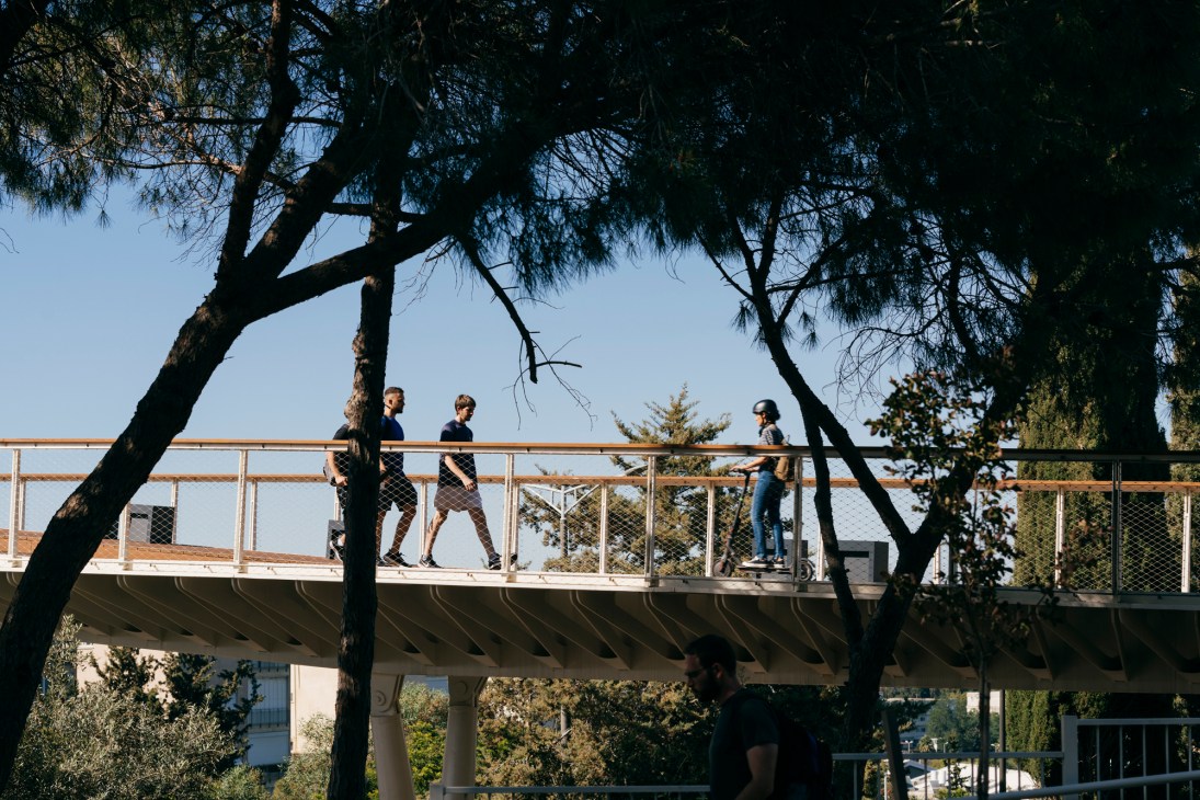 People walking across the Haifa Bridge pedestrian walkway with metal railings and trees framing the view.