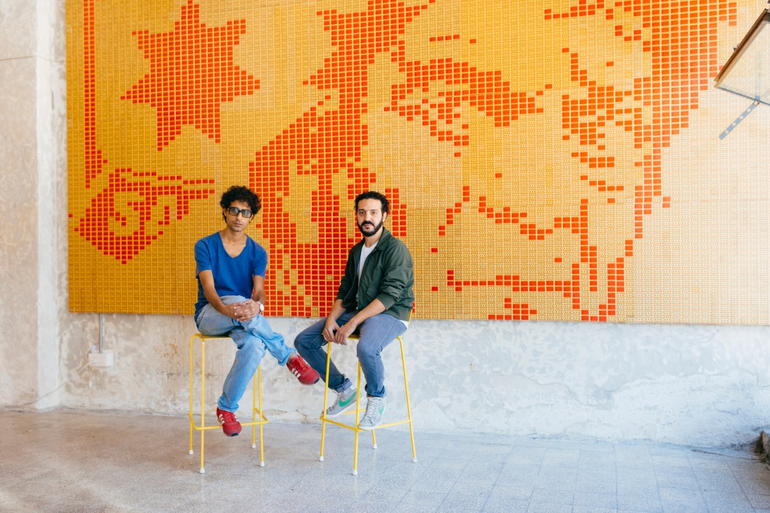 Two men sit on yellow chairs against a large orange and yellow pixelated wall mural in Amman.
