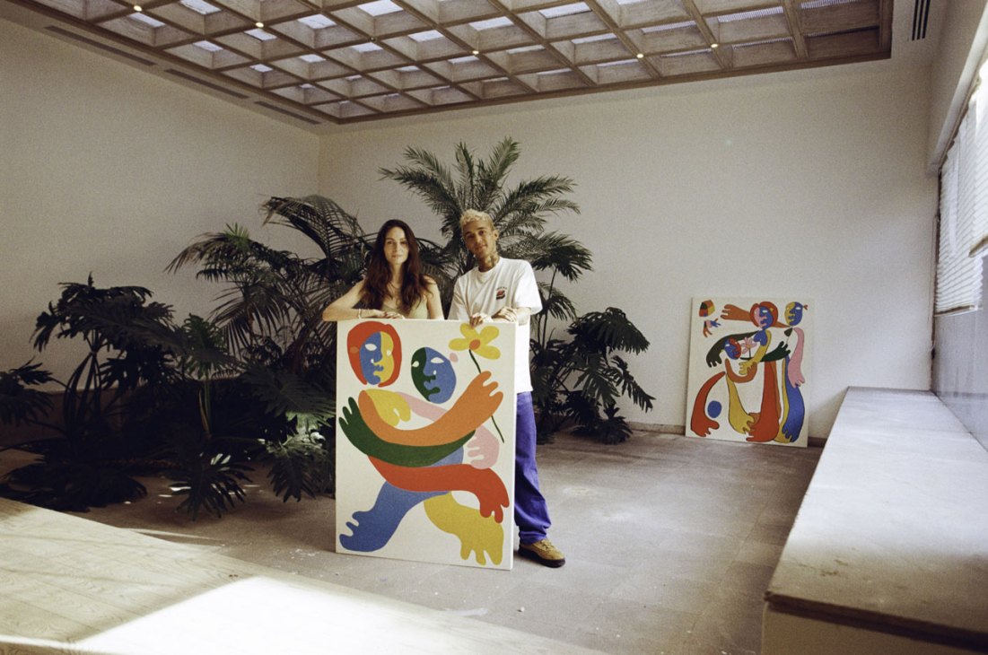 Two people posing with colorful abstract paintings in a modern interior space with plants and coffered ceiling