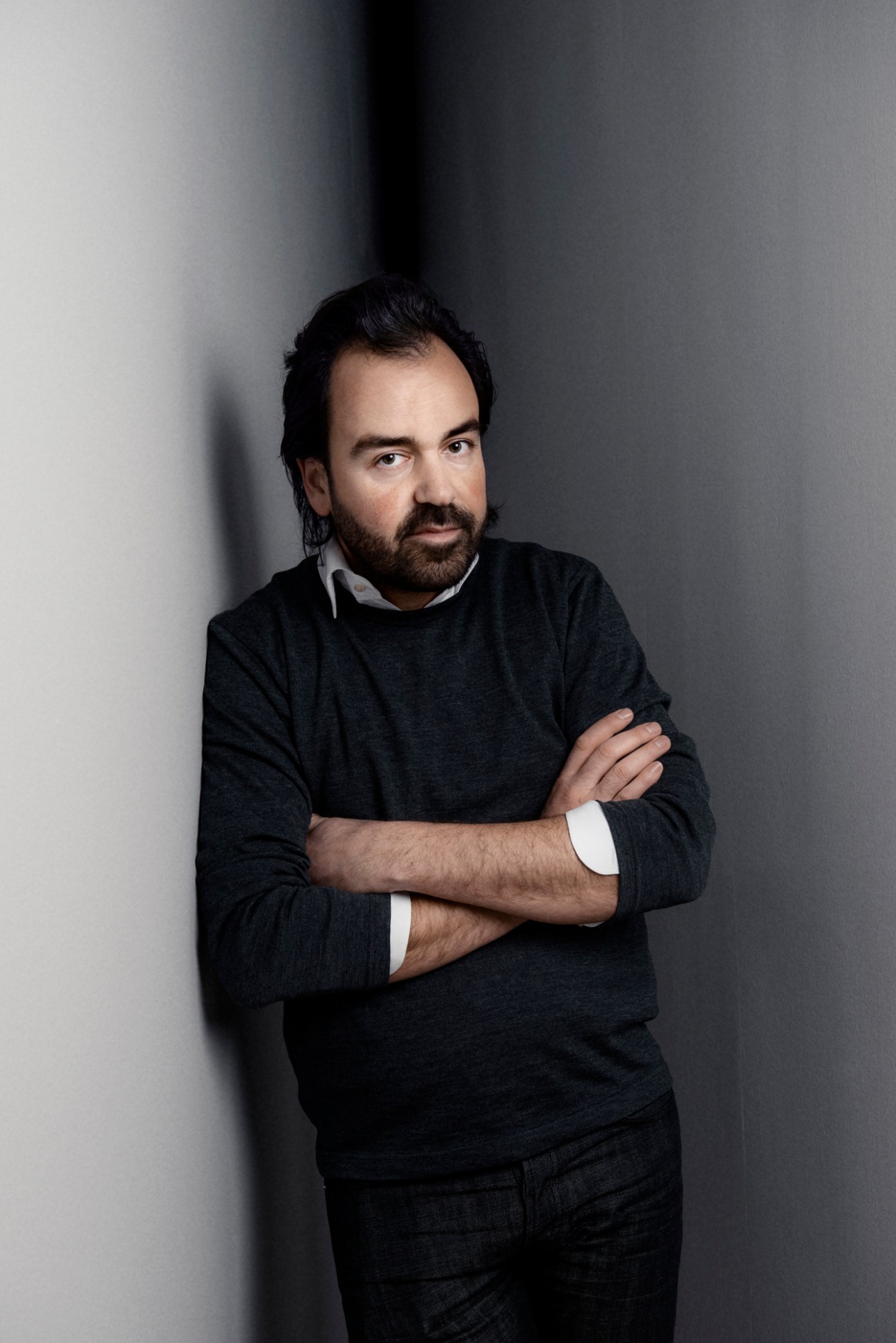 Man with dark hair and beard wearing dark sweater and collared shirt, arms crossed, against gray background