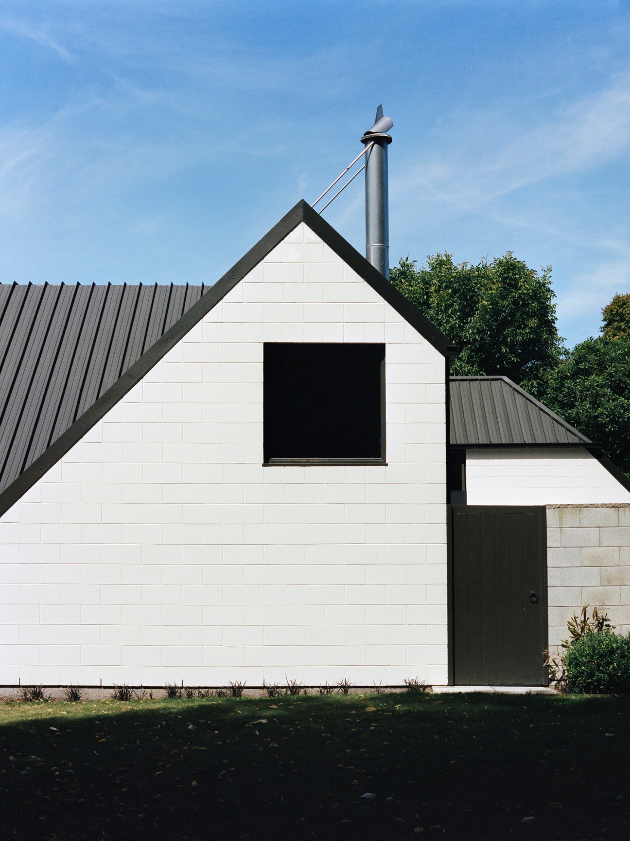White concrete block house with steep dark roof, black square window, and metal chimney against blue sky