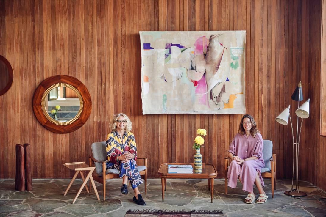 Two women sitting in mid-century modern chairs against a wood-paneled wall with abstract artwork and a circular mirror.