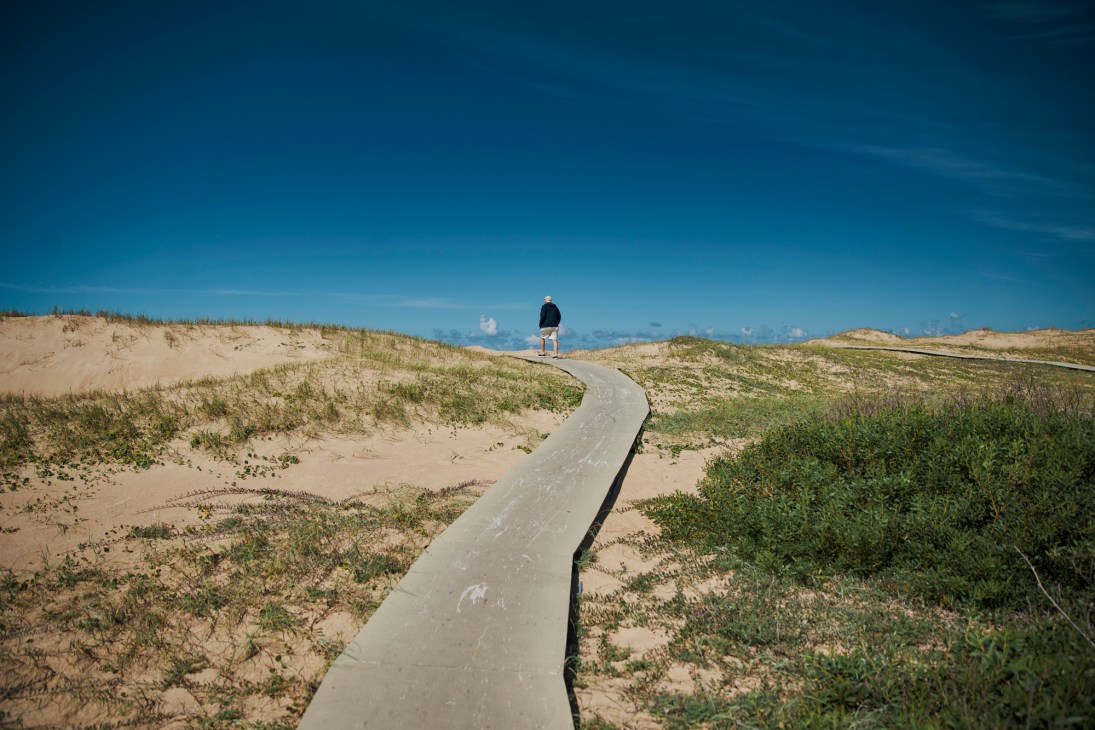 Person standing on a concrete path winding through sand dunes and beach vegetation.