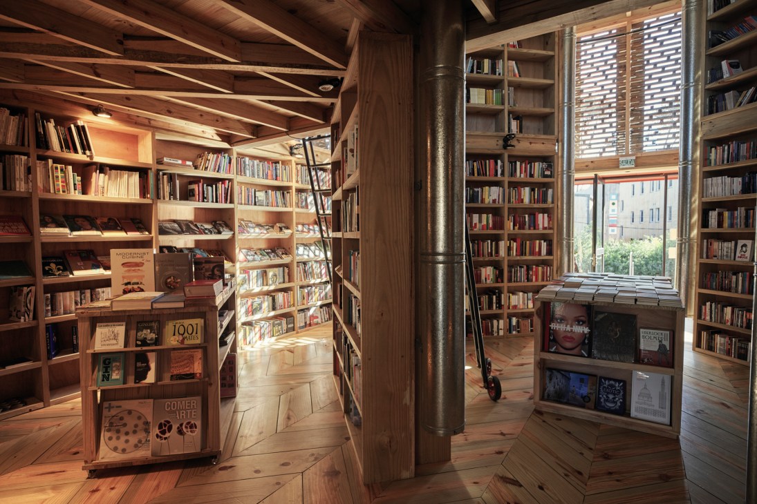 Wooden library shelves filled with books inside Rizoma bookshop, with large windows overlooking brick buildings.
