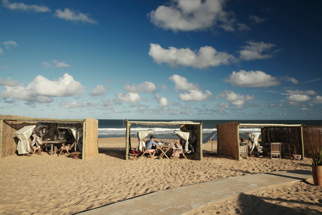 Diners eating at wooden tables under canvas canopies at Chiringuito beach restaurant with ocean waves visible behind them.