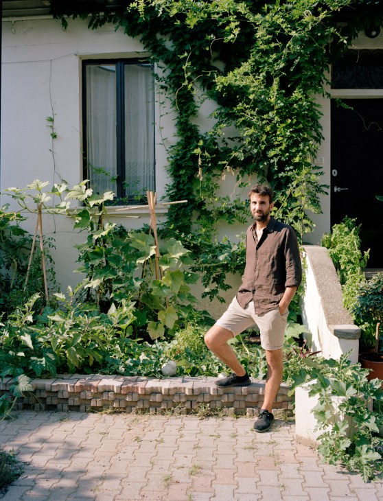 Joseph Grima stands in a lush garden courtyard surrounded by climbing ivy and green plants.