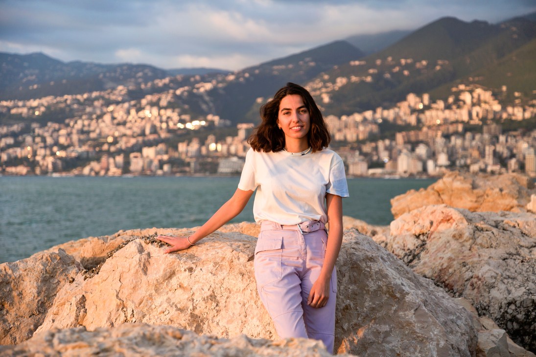 Dounia Messihi sitting on rocky coastline overlooking Mediterranean bay with hillside city in background