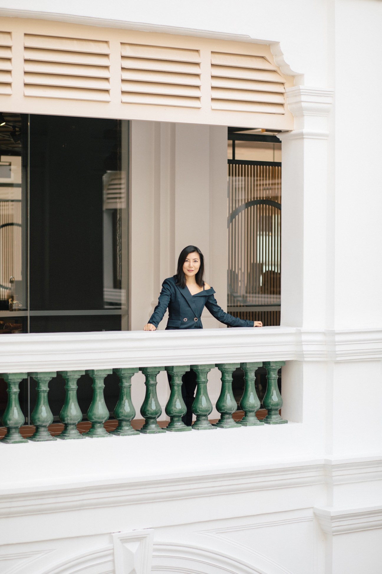 Woman in blue blazer leaning against a railing with green ornamental balusters on a white building facade.