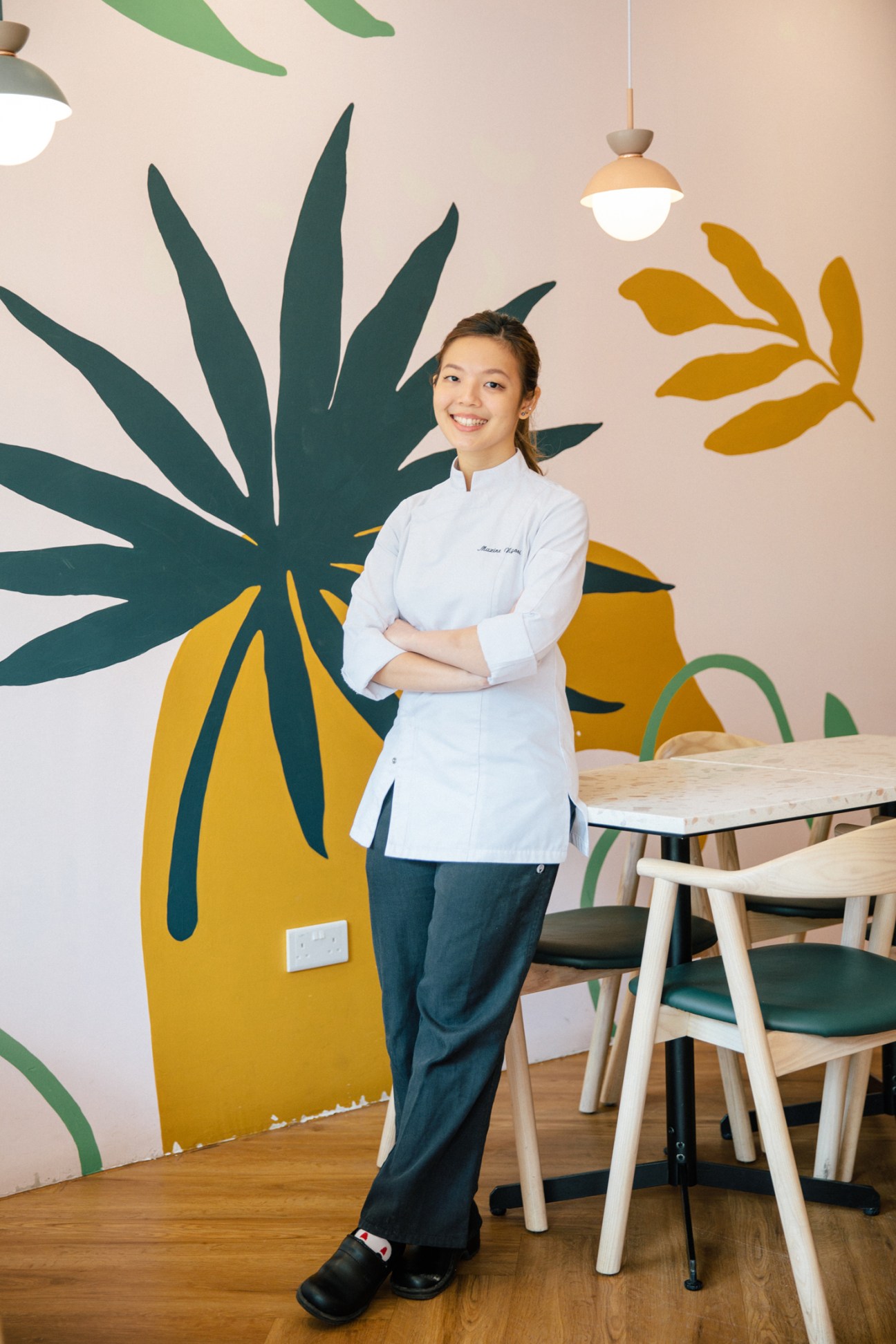 Smiling woman in white chef's jacket standing with arms crossed in colorful restaurant interior