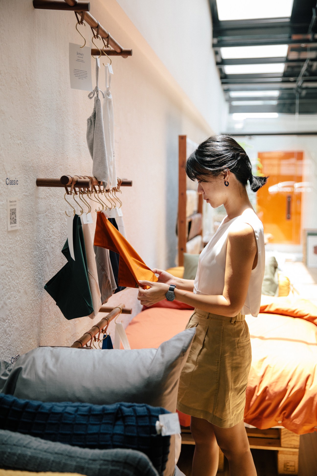 Woman in white top and tan skirt examining colorful fabrics hanging on wooden display rack