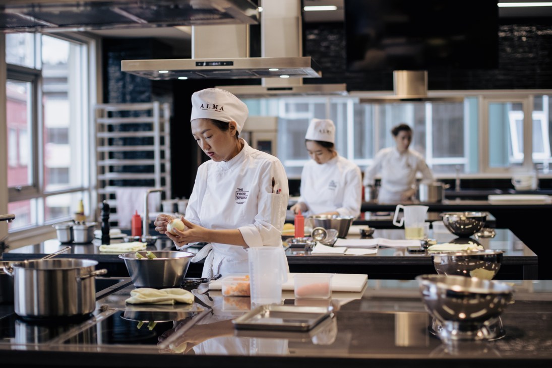 Chef instructing students in professional kitchen at The Food School in Bangkok, with students wearing white chef uniforms and ALMA branding.