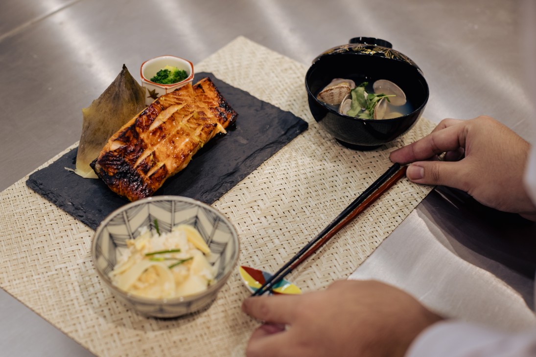 A person holding chopsticks over a Japanese meal plated on woven placemats with grilled fish, soup, and side dishes.