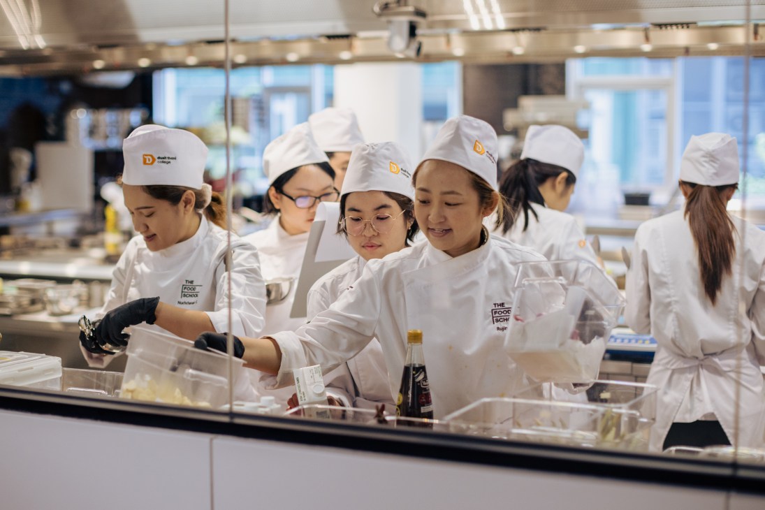 Students in white chef coats and hats work at cooking stations in a professional kitchen classroom at The Food School Bangkok.