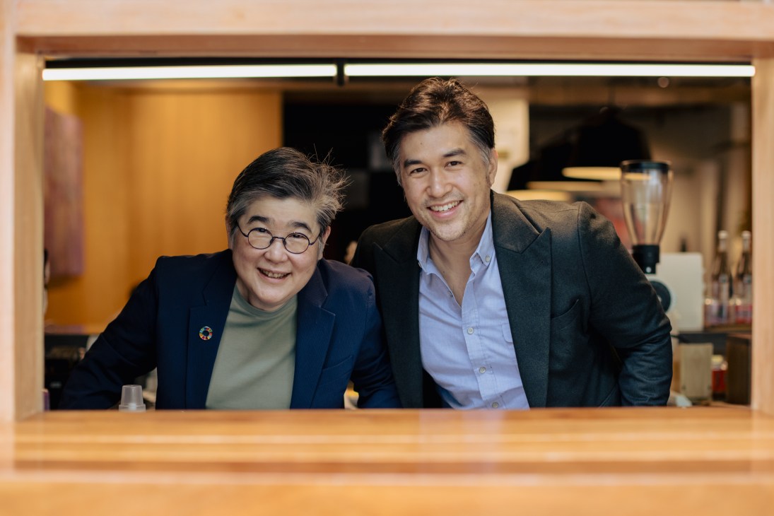 Two men smile at the camera from behind a wooden counter in a modern kitchen setting.