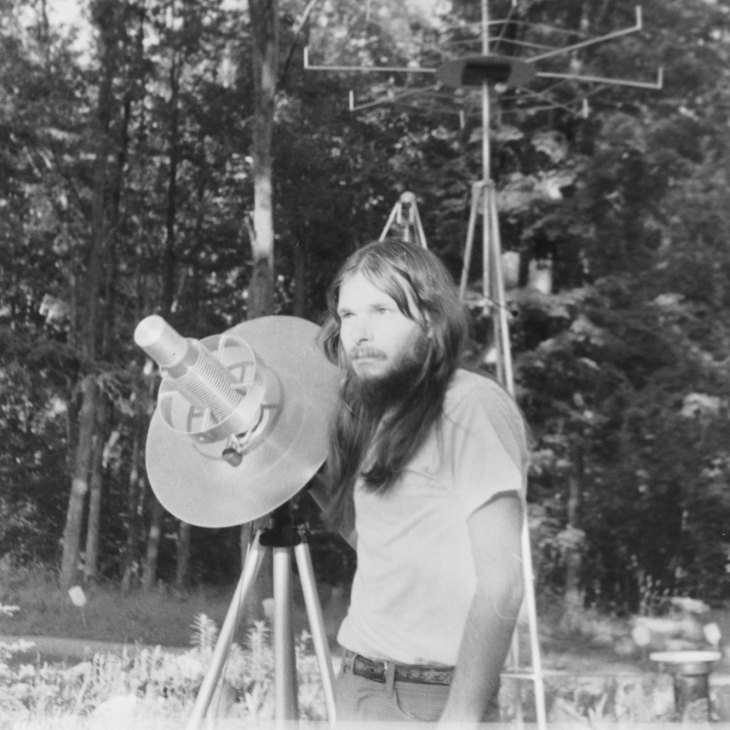 Man with long hair and beard using telescope equipment outdoors in black and white documentary still