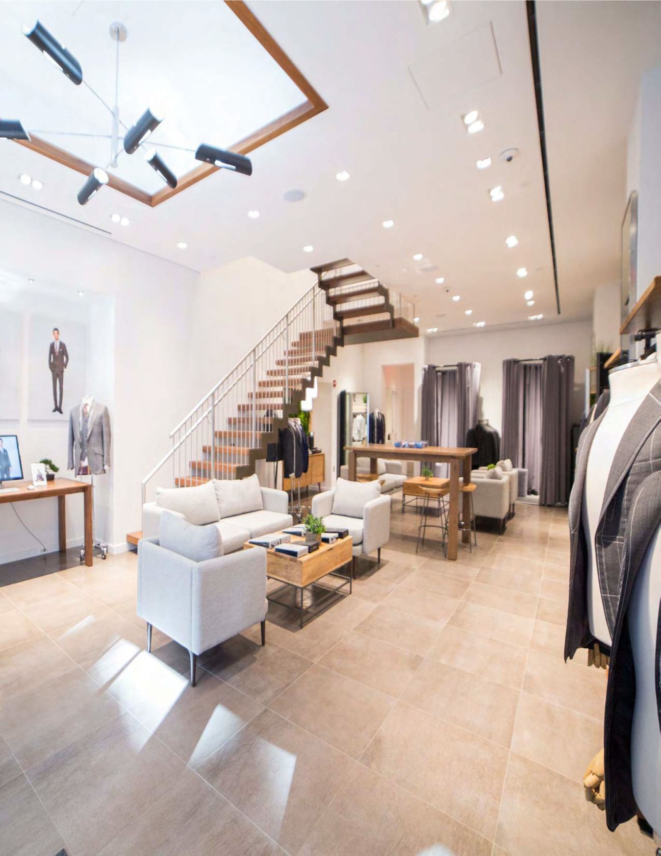 Modern menswear retail store interior with grey seating area, wooden staircase, and suits displayed on racks