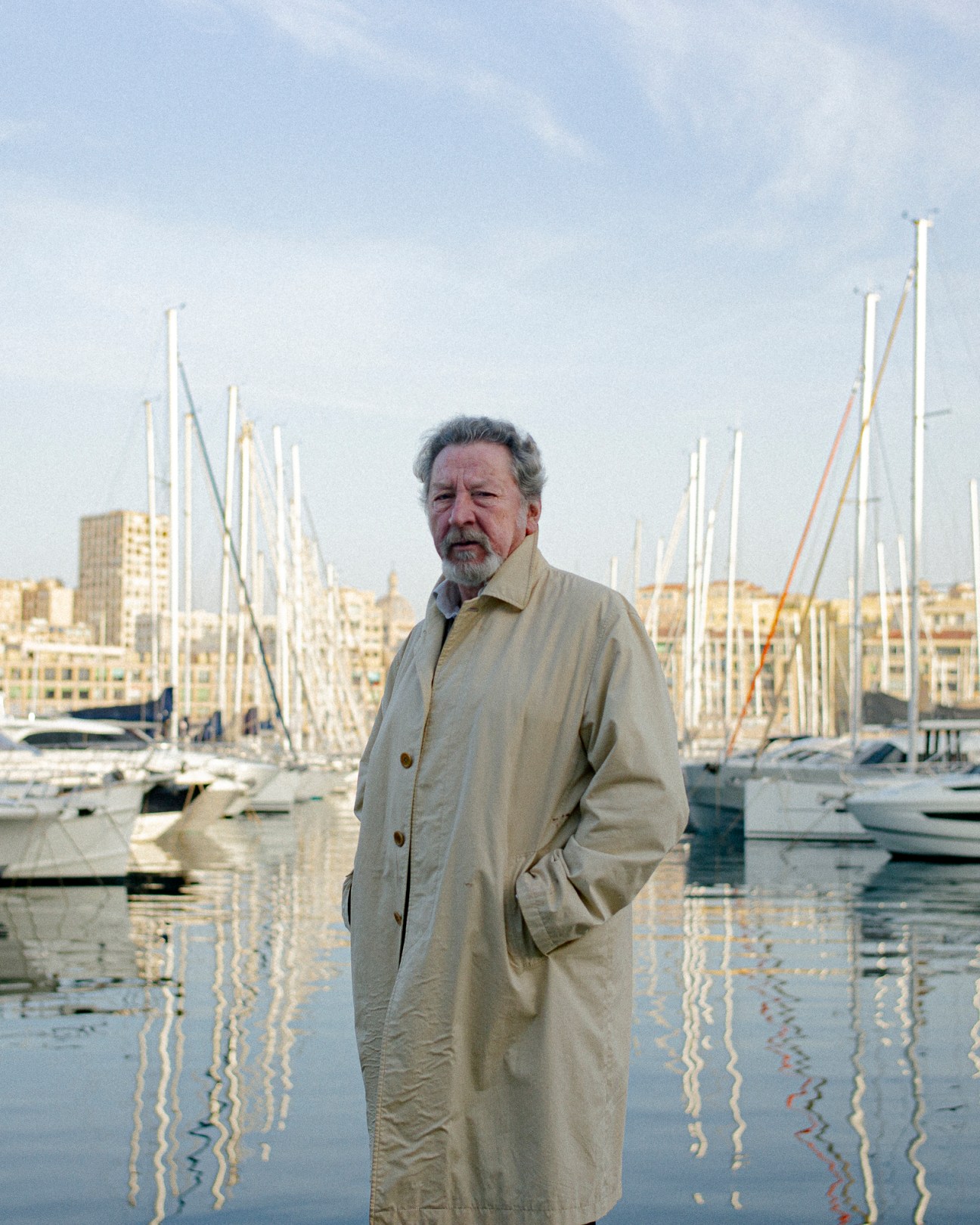 Director Robert Guédiguian stands in a beige coat at Marseille's old port with sailboat masts in background
