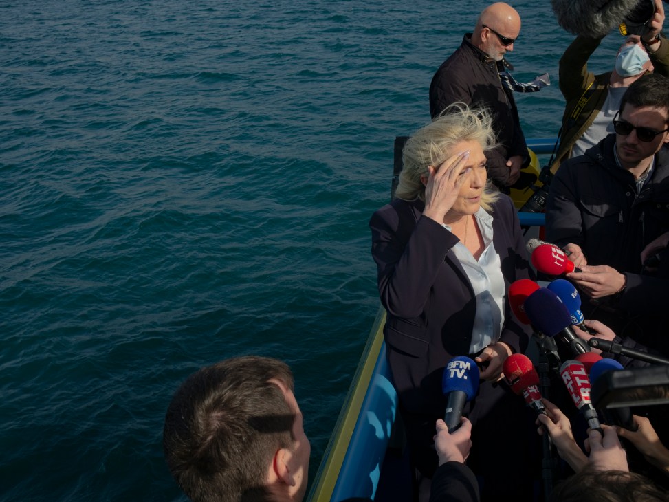 Blonde woman in dark blazer speaks to journalists holding microphones on boat deck with blue water behind
