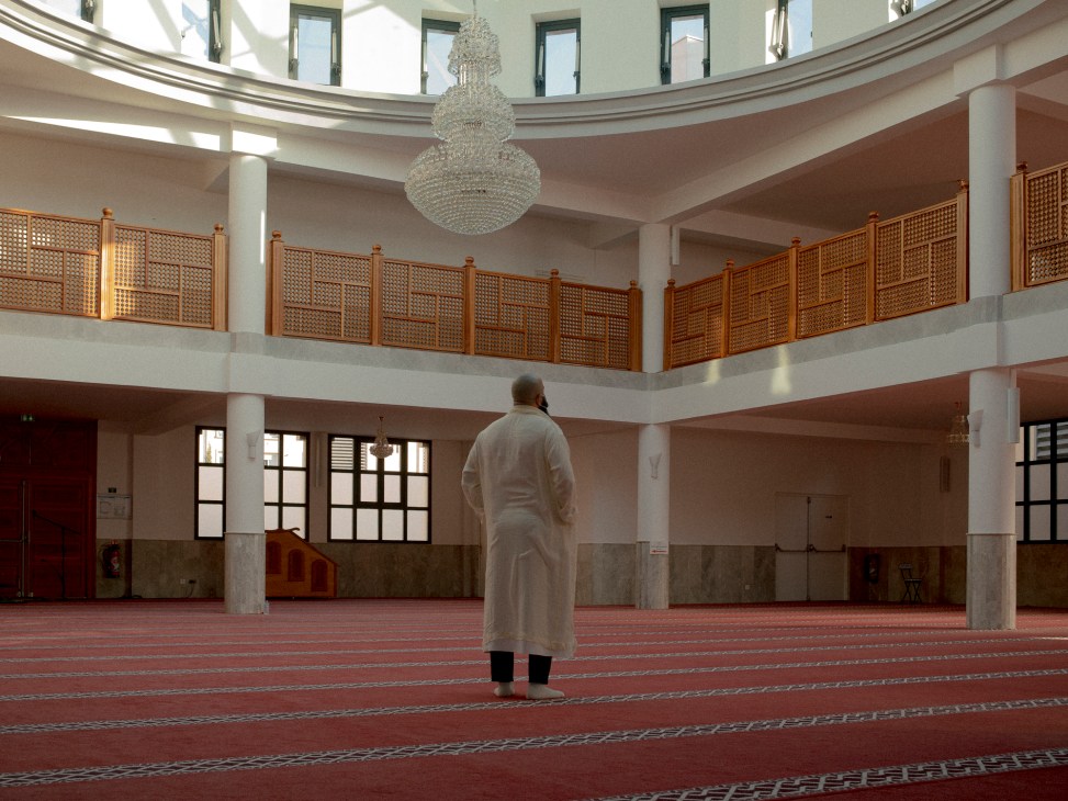 A man in white robes stands alone in prayer inside a modern mosque with ornate chandelier and red carpeting