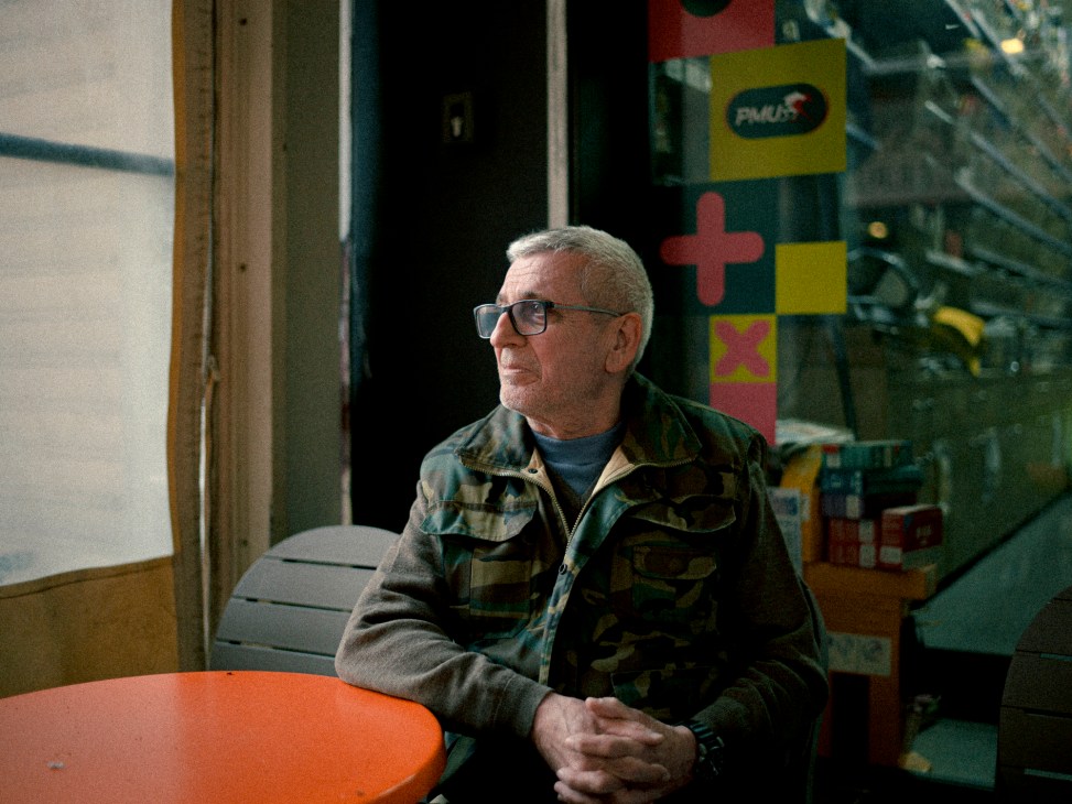 Elderly man in glasses and camouflage jacket sits at orange table in French cafe, looking thoughtfully to the side