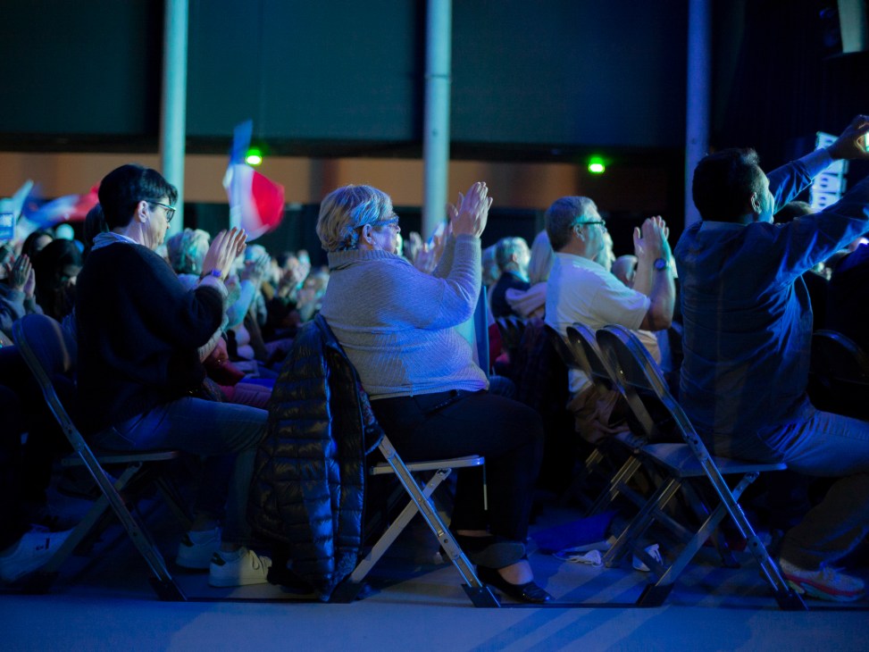 Audience members applauding at a political rally, seated on folding chairs under blue stage lighting