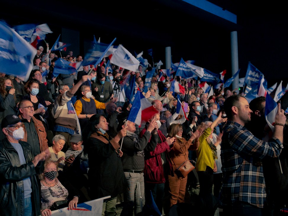 Crowd of supporters waving French flags at an indoor political rally, many wearing face masks