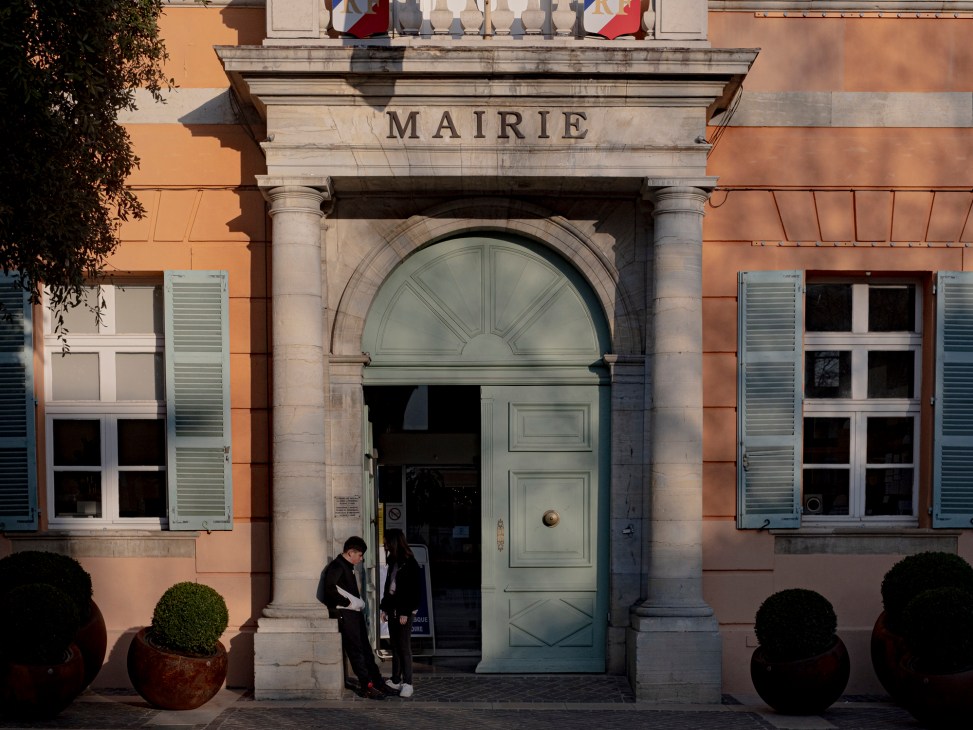 Two people stand outside the entrance of a French municipal building with 'MAIRIE' inscribed above stone columns