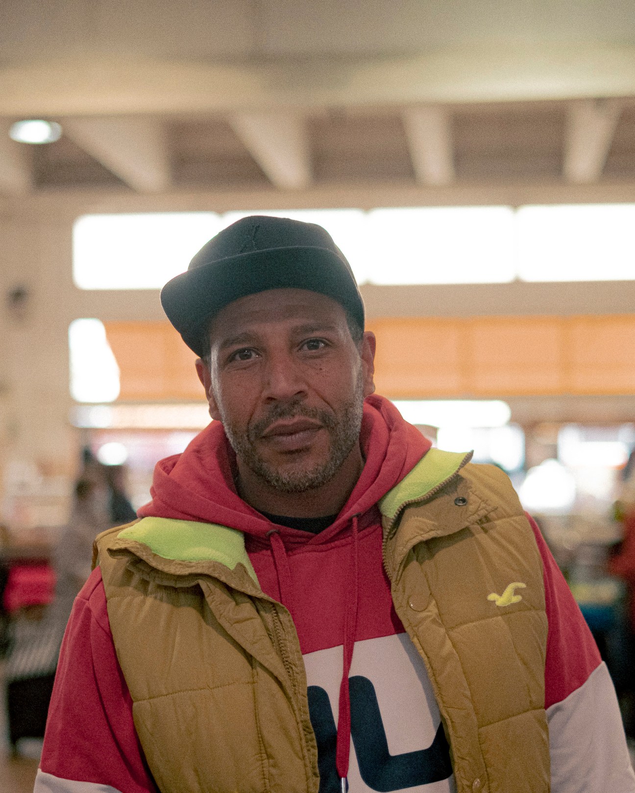 A middle-aged man in a dark beanie, red hoodie and tan jacket stands in what appears to be a hotel or restaurant interior