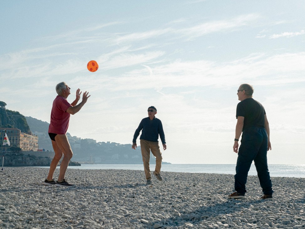 Three elderly men playing with an orange ball on a pebble beach with coastal buildings and water in background